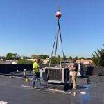 two men work to align A/C unit from crane on to roof